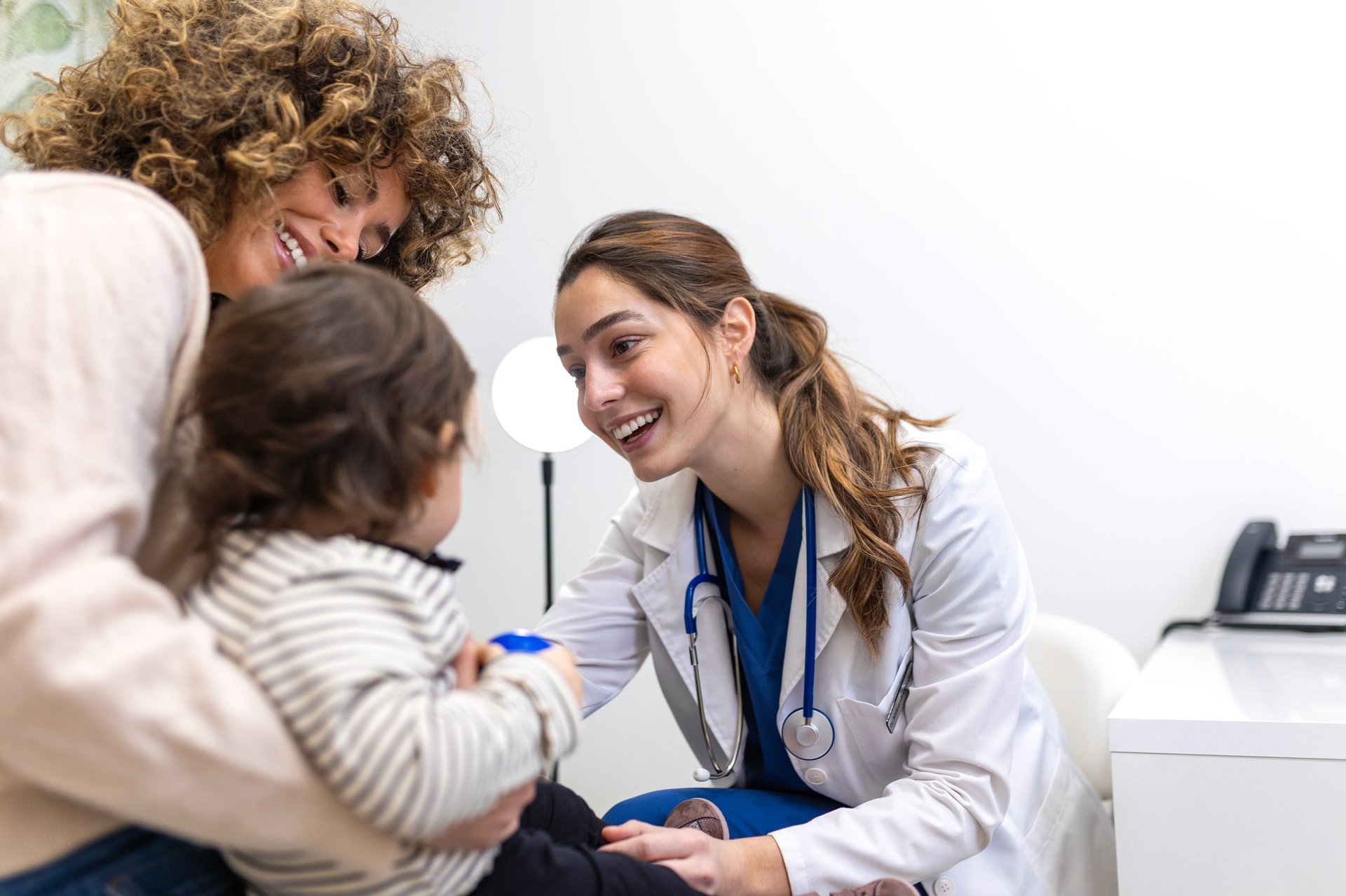 Pediatrician examining little child with friendly female doctor smiling at toddler during medical checkup