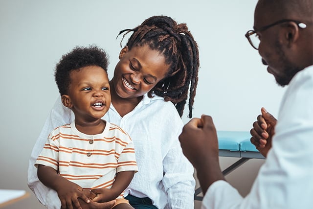 A woman holding a smiling toddler while interacting with a man in a medical setting